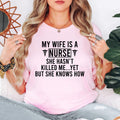 My Wife Is A Nurse She Hasn't Killed Me Yet But She Knows How T Shirt featuring a funny nursing quote designed for husbands of nurses, registered nurses, healthcare workers, and medical professionals. Teezora Prints creates premium nurse apparel for customers searching nurse husband shirts, RN clothing, funny nursing gifts, hospital staff apparel, healthcare fashion, medical graphic tees, and unique nurse appreciation gifts.