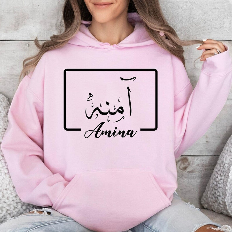 Elegant personalised Arabic name hoodie showcasing a premium gold Arabic calligraphy design displayed within a modern geometric frame. The minimalist composition blends traditional Arabic script with contemporary typography to create a sophisticated and eye-catching appearance. Printed on a black hoodie for maximum visual contrast, the luxurious gold lettering enhances the overall design while maintaining a clean and timeless aesthetic suitable for all ages.