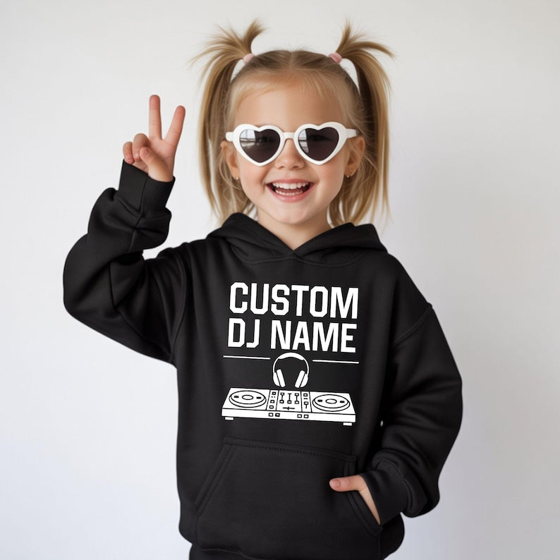 Personalised Kids DJ Hoodie featuring a custom DJ name with a modern controller and headphones graphic, designed for young music lovers who dream of becoming DJs. Perfect for birthdays, family outings, school holidays, music events and everyday comfort. Every premium hoodie is professionally printed in the UK by TeezoraPrints using soft, durable fabric for all-day wear.