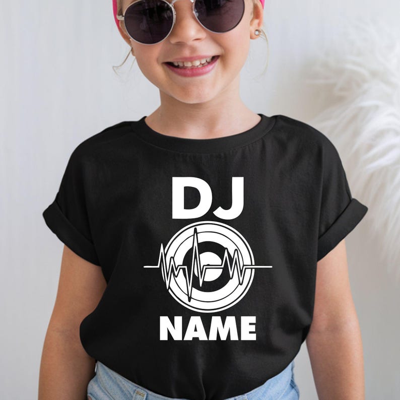 A child wearing a black short-sleeve T-shirt with a white 'DJ NAME' graphic on the front, paired with light blue jeans and round sunglasses, against a light indoor background.