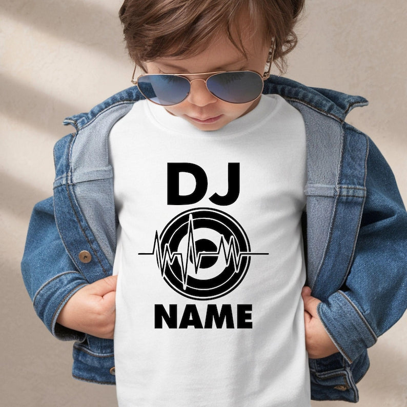A child wearing aviator sunglasses, a blue denim jacket, and a white t-shirt with a black graphic that reads 'DJ NAME' with a circular heartbeat-style logo. The child is posed against a neutral beige background.