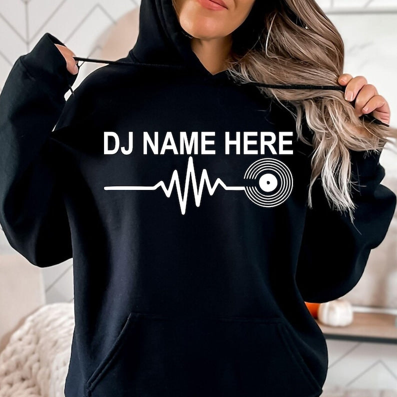 Personalised DJ Name Heartbeat Hoodie featuring a custom DJ name, heartbeat waveform and vinyl record graphic. Designed for DJs, music producers, club performers and electronic music lovers, this premium unisex hoodie is professionally printed in the UK by TeezoraPrints for warmth, comfort and everyday style.