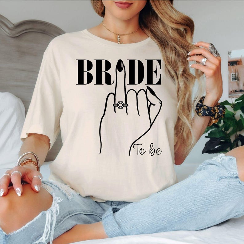 Looking for a thoughtful gift for the future bride? This Bride To Be shirt is a perfect engagement gift for fiancées, brides-to-be and newly engaged women. Featuring an elegant bridal illustration and modern design, every shirt is professionally printed in the UK by TeezoraPrints, making it a memorable keepsake for every wedding celebration.