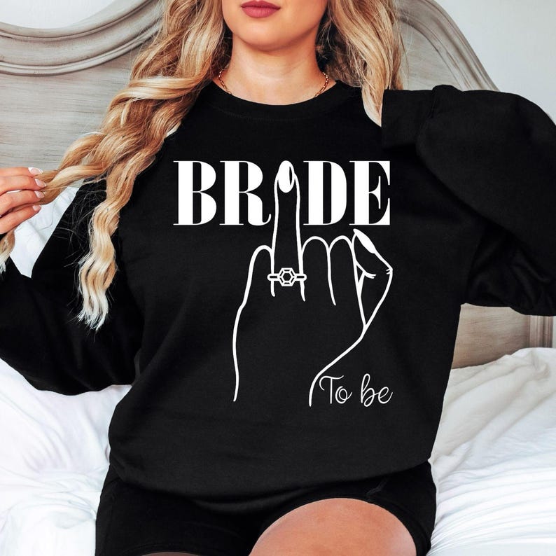 Personalised Bride To Be sweatshirt designed for brides, fiancées and future wives celebrating their engagement and upcoming wedding. This elegant sweatshirt features a minimalist bridal illustration with an engagement ring, creating a timeless look for every bride. Perfect for staying cosy during wedding planning, with every sweatshirt professionally printed in the UK by TeezoraPrints using premium quality materials.