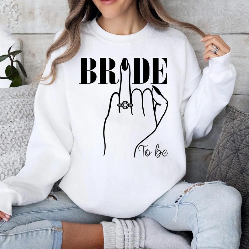 Personalised Bride To Be sweatshirt designed for brides, fiancées and future wives celebrating their engagement and upcoming wedding. This elegant sweatshirt features a minimalist bridal illustration with an engagement ring, creating a timeless look for every bride. Perfect for staying cosy during wedding planning, with every sweatshirt professionally printed in the UK by TeezoraPrints using premium quality materials.