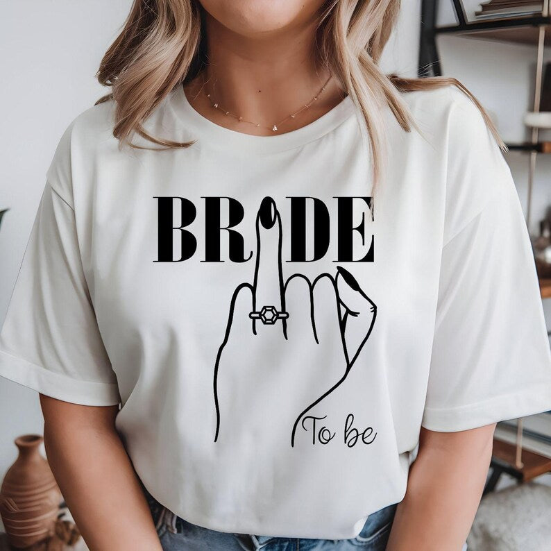 Bride To Be shirt designed for brides, fiancées and future wives celebrating their engagement and wedding journey. Featuring a stylish bridal illustration with an engagement ring and elegant "To Be" wording, this premium bridal t-shirt is perfect for women looking for a fashionable outfit for pre-wedding celebrations. Professionally printed in the UK by TeezoraPrints using premium quality materials for comfort and durability.