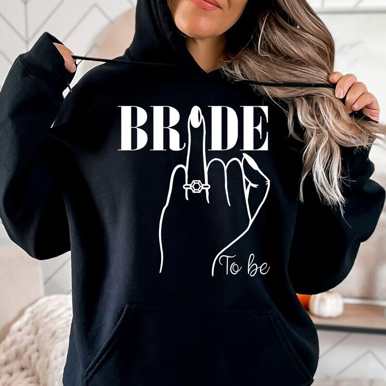 Personalised Bride To Be hoodie created for brides, fiancées and future wives celebrating one of life's most memorable milestones. Featuring an elegant bride illustration with a sparkling engagement ring and stylish "To Be" design, this cosy hoodie is perfect for staying warm while planning your wedding. Every hoodie is professionally printed in the UK by TeezoraPrints using premium-quality materials for lasting comfort.