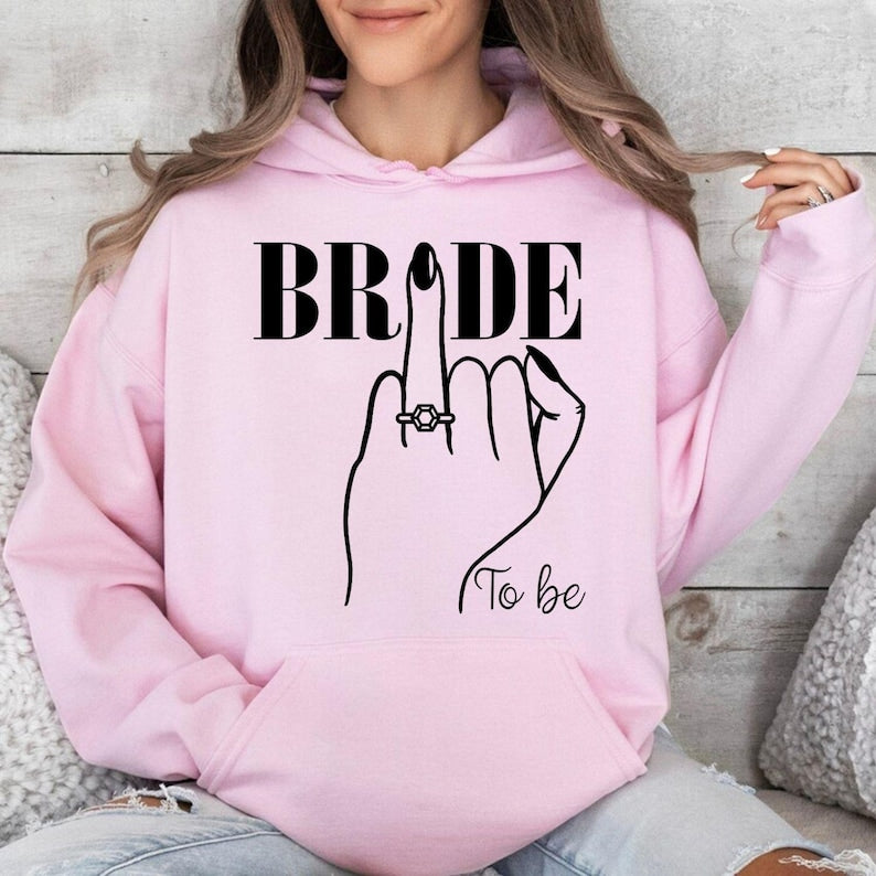 Personalised Bride To Be hoodie created for brides, fiancées and future wives celebrating one of life's most memorable milestones. Featuring an elegant bride illustration with a sparkling engagement ring and stylish "To Be" design, this cosy hoodie is perfect for staying warm while planning your wedding. Every hoodie is professionally printed in the UK by TeezoraPrints using premium-quality materials for lasting comfort.