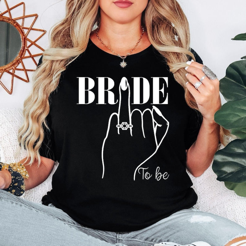 Bride To Be shirt designed for brides, fiancées and future wives celebrating their engagement and wedding journey. Featuring a stylish bridal illustration with an engagement ring and elegant "To Be" wording, this premium bridal t-shirt is perfect for women looking for a fashionable outfit for pre-wedding celebrations. Professionally printed in the UK by TeezoraPrints using premium quality materials for comfort and durability.