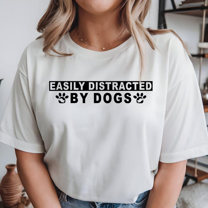 Easily Distracted By Dogs t shirt created for dog lovers, dog mums, dog dads, puppy parents, pet owners, animal rescuers, dog walkers, veterinarians, groomers, trainers, and anyone who instantly smiles whenever they see a dog. Featuring a bold typography design with cute paw prints, this premium graphic tee celebrates the special bond between people and their furry companions while delivering exceptional comfort, stylish everyday fashion, and a fun personality that every dog enthusiast will instantly relate