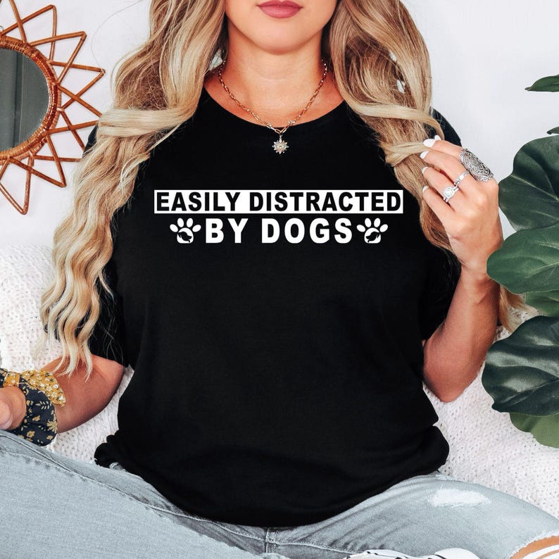 Easily Distracted By Dogs t shirt created for dog lovers, dog mums, dog dads, puppy parents, pet owners, animal rescuers, dog walkers, veterinarians, groomers, trainers, and anyone who instantly smiles whenever they see a dog. Featuring a bold typography design with cute paw prints, this premium graphic tee celebrates the special bond between people and their furry companions while delivering exceptional comfort, stylish everyday fashion, and a fun personality that every dog enthusiast will instantly relate