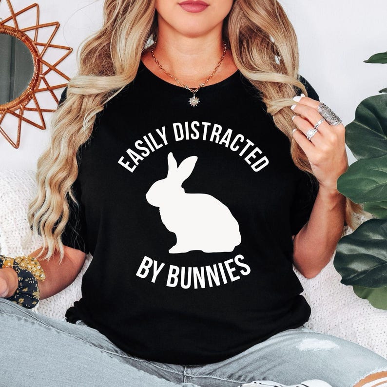 Easily Distracted By Bunnies T Shirt featuring a minimalist rabbit silhouette designed for rabbit owners, bunny lovers, pet parents, and animal enthusiasts. Teezora Prints combines premium quality with modern typography, making this shirt perfect for customers searching rabbit shirts, bunny clothing, funny bunny tees, rabbit owner gifts, pet lover apparel, bunny graphic t shirts, premium bunny clothing, unique animal lover apparel, and everyday casual bunny-themed outfits.