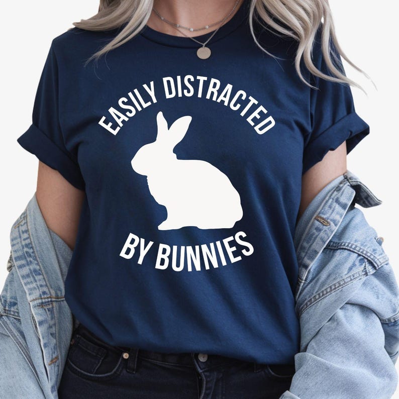 Easily Distracted By Bunnies T Shirt created for bunny lovers, rabbit owners, pet parents, animal enthusiasts, rabbit breeders, rescue volunteers, farm lovers, veterinary staff, and anyone who instantly smiles whenever they see a bunny. Featuring a bold rabbit silhouette with clean typography, this premium graphic tee from Teezora Prints celebrates a love for adorable rabbits while delivering exceptional comfort, premium print quality, stylish everyday fashion, and a fun personality for every bunny enthusia