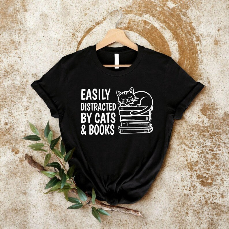 Easily Distracted By Cats & Books T Shirt created for cat lovers, bookworms, avid readers, librarians, teachers, students, writers, novel enthusiasts, pet parents, and anyone who enjoys reading with a cat by their side. Featuring a sleepy cat resting on stacked books with bold typography, this premium graphic tee from Teezora Prints celebrates two favourite passions while offering exceptional comfort, premium print quality, stylish everyday fashion, and timeless book lover appeal.