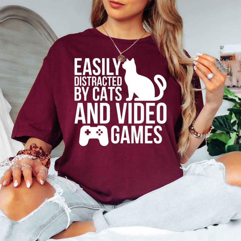 Easily Distracted By Cats And Video Games T Shirt featuring a funny gaming quote with a cat silhouette and controller graphic designed for gamers and cat lovers. Teezora Prints creates premium apparel for customers searching gamer shirts, funny gaming tees, cat owner clothing, gaming gifts, gaming apparel, PC gamer t shirts, console gamer clothing, esports fashion, cat lover gifts, premium graphic tees, and unique gaming lifestyle clothing.