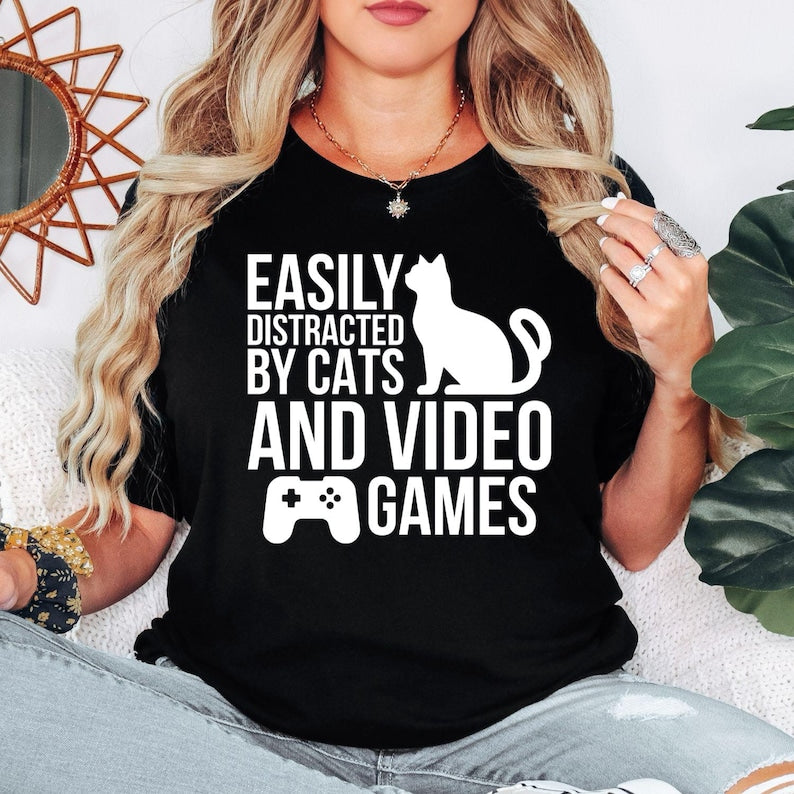 Easily Distracted By Cats And Video Games T Shirt created for cat lovers, gamers, streamers, console players, PC gamers, pet parents, esports fans, and anyone whose two greatest passions are cats and gaming. Featuring a playful cat silhouette with a game controller, this premium graphic tee from Teezora Prints delivers exceptional comfort, professional print quality, stylish everyday fashion, and the perfect conversation starter for gaming and feline enthusiasts alike.