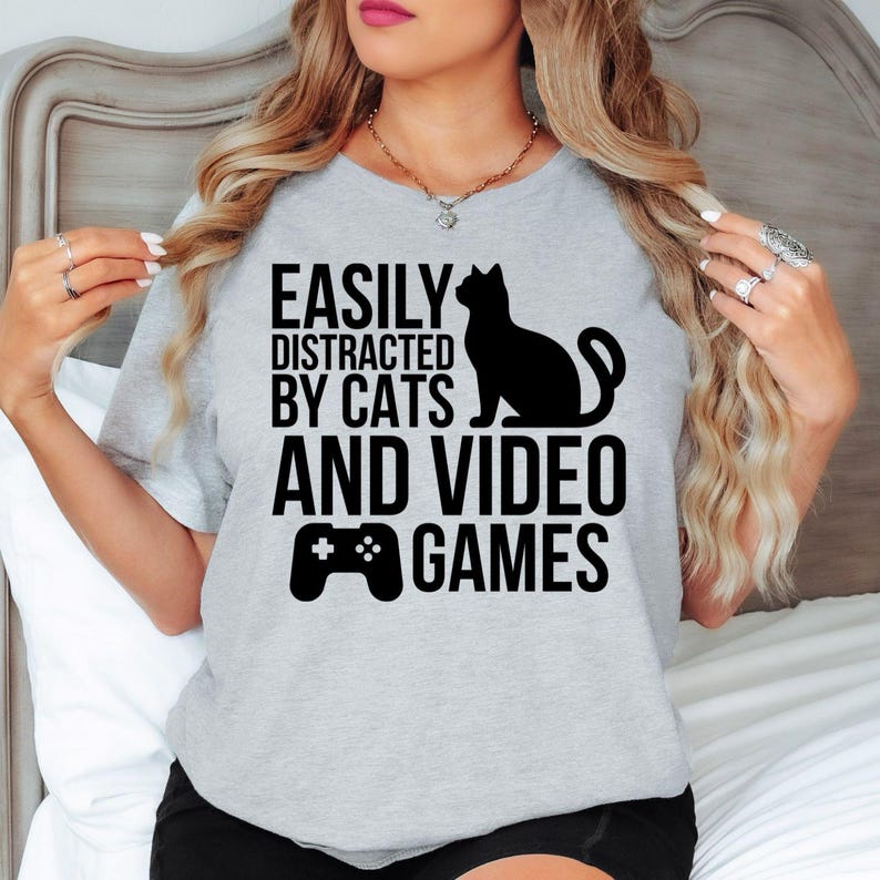 Easily Distracted By Cats And Video Games T Shirt makes an unforgettable gift for gamers, cat mums, cat dads, streamers, Twitch creators, YouTubers, esports players, brothers, sisters, sons, daughters, husbands, wives, friends, coworkers, and pet lovers. Designed by Teezora Prints, this premium graphic shirt is perfect for birthdays, Christmas, Father's Day, Mother's Day, gaming events, Secret Santa, and thoughtful gifts for anyone obsessed with cats and video games.