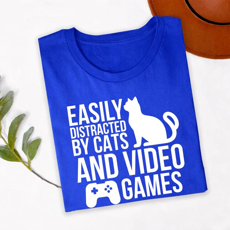 Easily Distracted By Cats And Video Games T Shirt is made for people whose ideal day includes playing video games while spending time with their favourite cat. Whether you're grinding ranked matches, relaxing with indie games, watching esports, or cuddling your pet after a gaming session, this premium graphic tee from Teezora Prints perfectly represents your personality. Featuring a bold cat silhouette and gaming controller design, it's ideal for everyday wear, travel, gaming events, holidays, and casual st