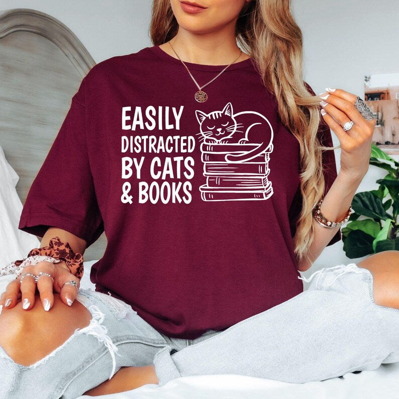Easily Distracted By Cats & Books T Shirt featuring a cute sleeping cat on stacked books designed for bookworms, readers, librarians, teachers, students, and cat lovers. Teezora Prints creates premium apparel for customers searching book lover shirts, cat lover clothing, reading t shirts, library apparel, bookworm gifts, literary fashion, reading quote shirts, cat graphic tees, premium book-themed clothing, and unique gifts for passionate readers.