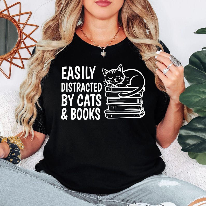 Easily Distracted By Cats & Books T Shirt created for cat lovers, bookworms, avid readers, librarians, teachers, students, writers, novel enthusiasts, pet parents, and anyone who enjoys reading with a cat by their side. Featuring a sleepy cat resting on stacked books with bold typography, this premium graphic tee from Teezora Prints celebrates two favourite passions while offering exceptional comfort, premium print quality, stylish everyday fashion, and timeless book lover appeal.
