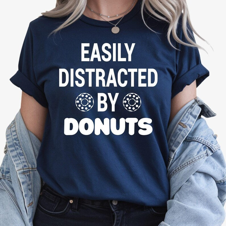 Show everyone your love for sweet treats with this Easily Distracted by Donuts graphic t-shirt featuring bold typography and fun donut icons. The soft premium cotton and relaxed unisex fit make it perfect for everyday wear. Every order is made to order and professionally printed in the UK by TeezoraPrints with exceptional attention to detail.